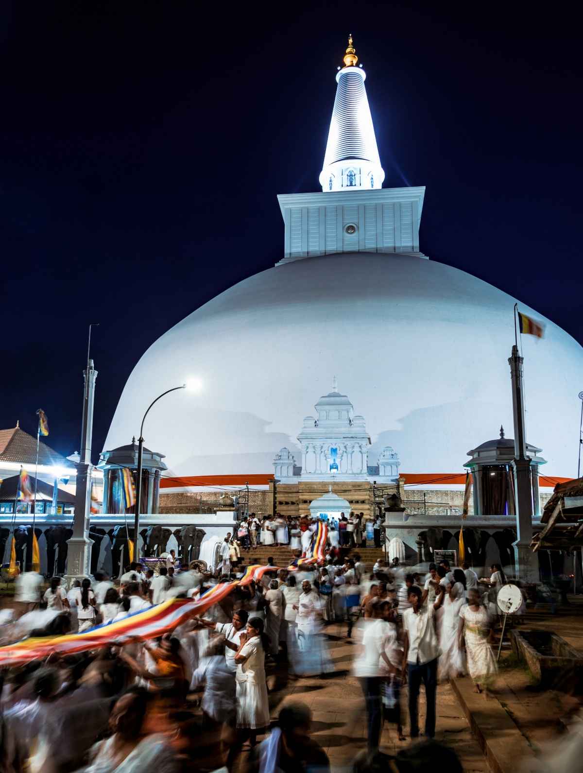 Photo of white stupa in nighttime with people celebrating fullmoon ceremonies carrying hundreds of meters long linen towards the temple to honour Buddha.